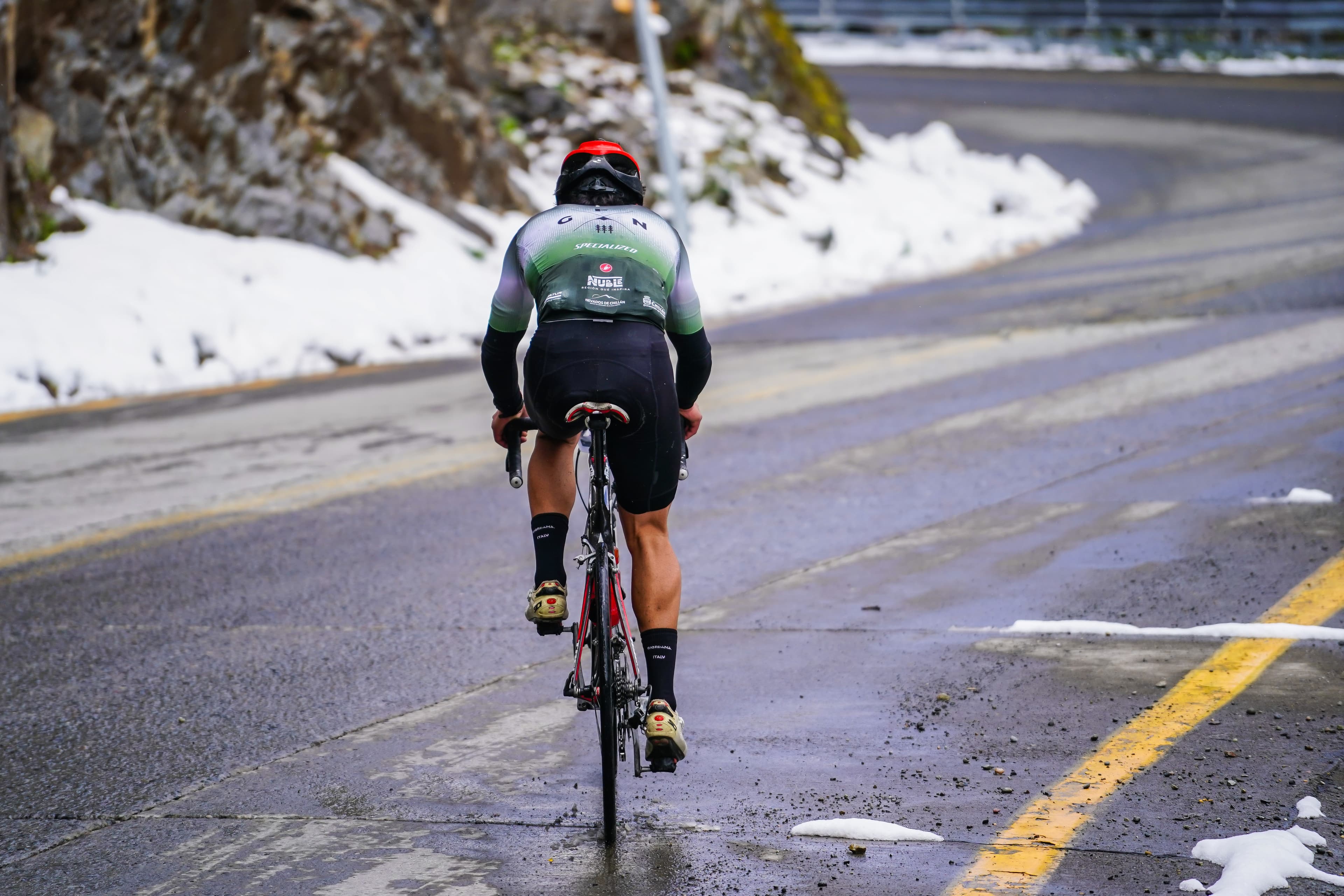 Ciclista de espaldas en una carretera de montaña que curva hacia la derecha, con nieve a ambos costados.